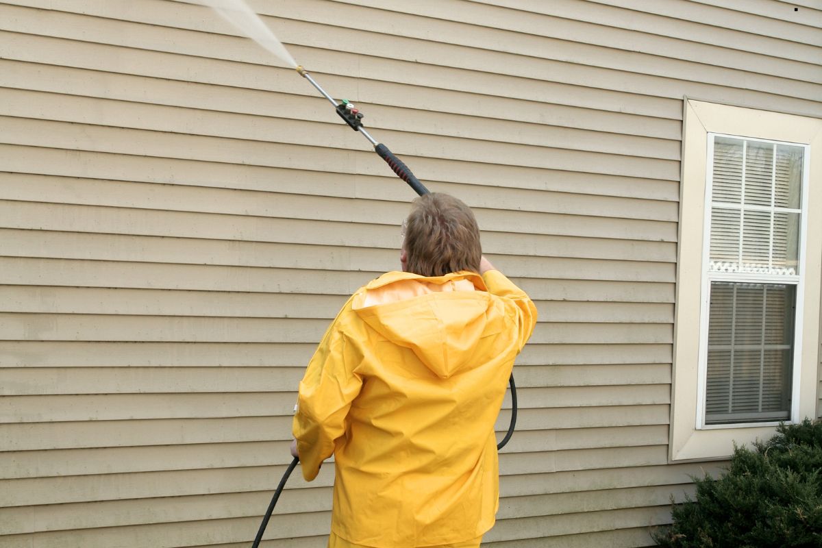 A man in a yellow raincoat operates a pressure washer, providing window cleaning services outdoors.