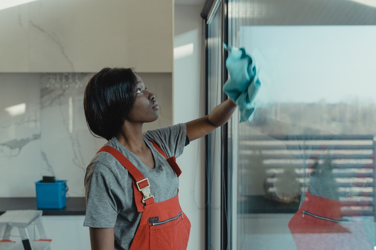 A woman wearing overalls is cleaning a window with a cloth, focused on her task in a bright, well-lit environment. 